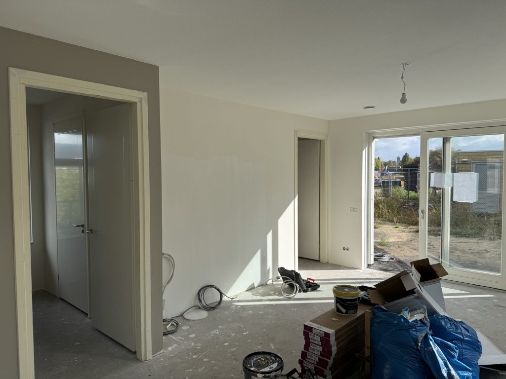 Woonkamer van een net opgeleverd huis met kale wanden, tuindeuren en bouwmaterialen op de vloer