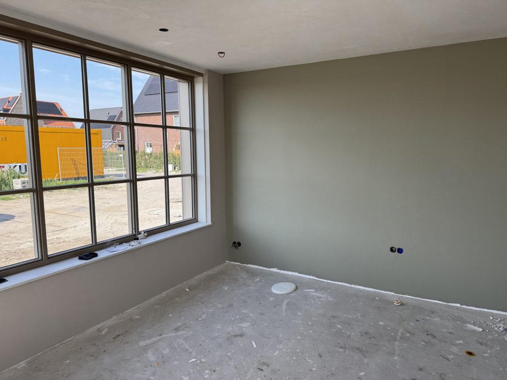 Nieuwbouwkamer met een groengrijs gesausde wand en grote ramen die uitkijken op een bouwterrein