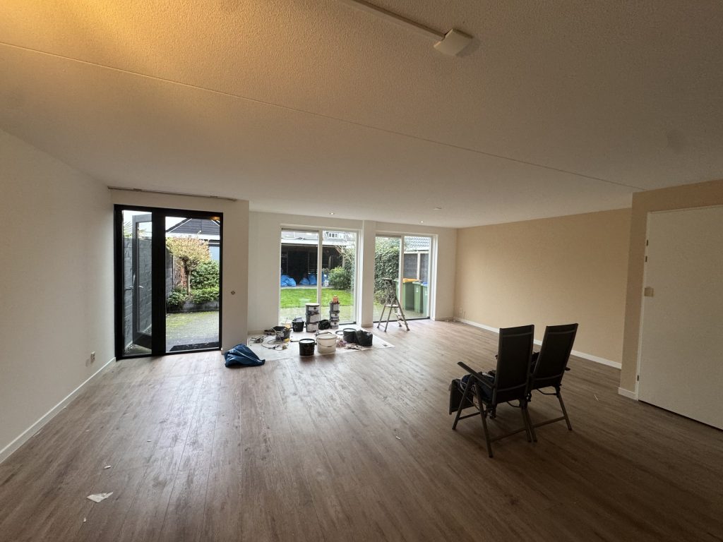 Leeggemaakte woonkamer in een bestaand huis, klaar voor nieuw behang, met verf en een trap bij de tuindeuren