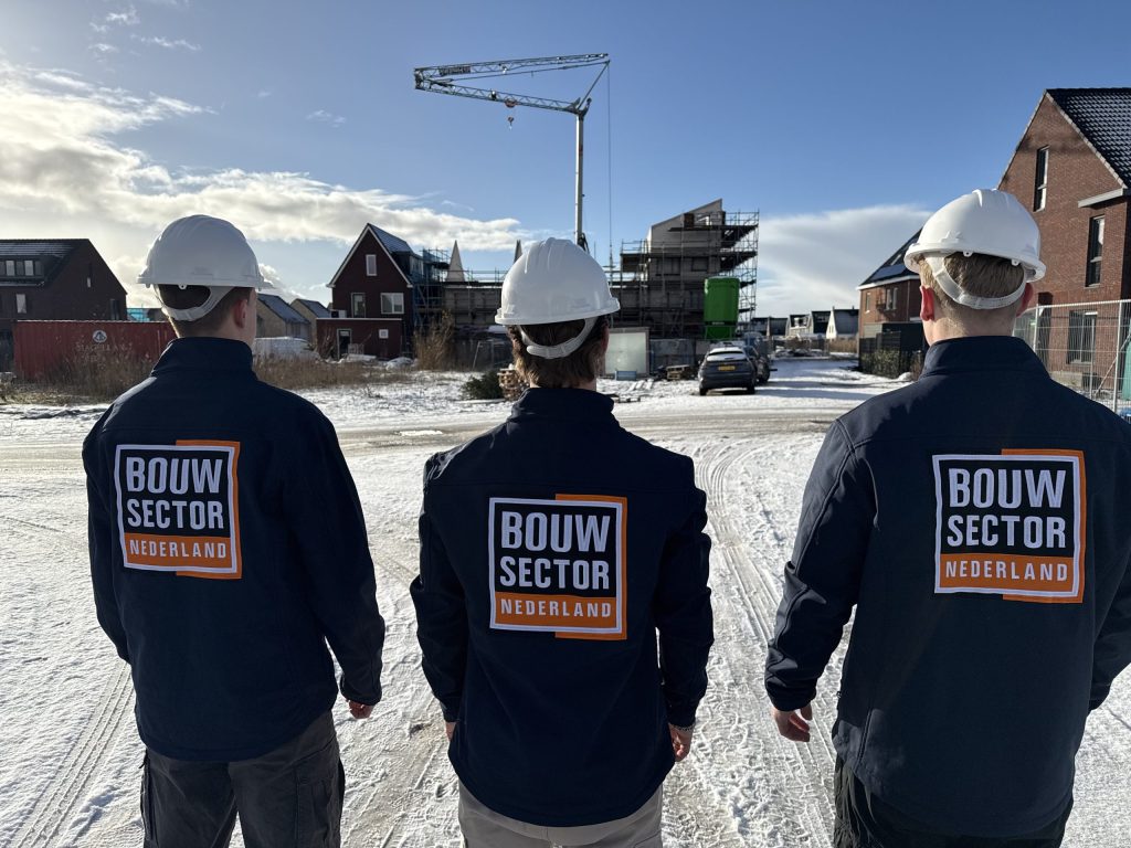 Drie vakmensen van Bouwsector Nederland met witte helm op een besneeuwde straat met nieuwbouw en een bouwkraan